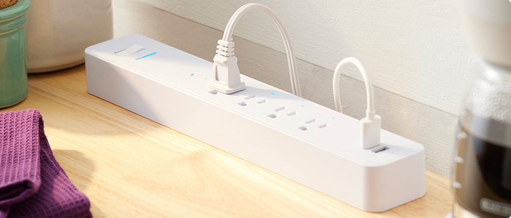 A white NOMA iQTM power bar rests on a hardwood floor inside a home. A white two-prong power cable and a white USB cable are plugged into the power bar.