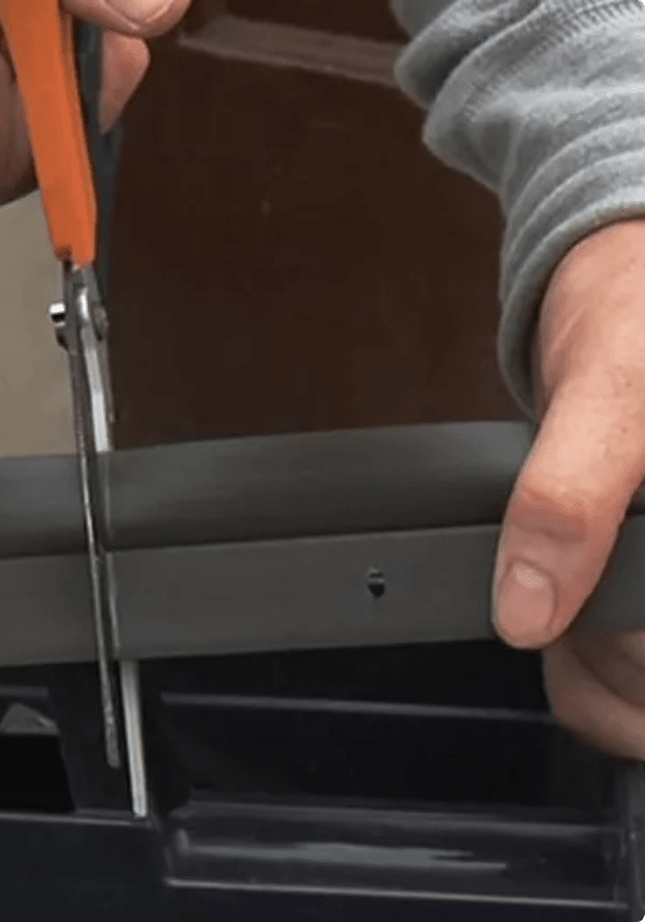 A hand cuts a length of black weatherstripping with a pair of orange-handled scissors.