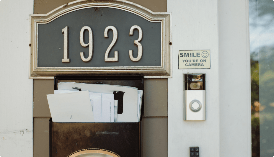 The front entrance to a home. A grey plaque with house numbers that read “1923” and a metal mailbox stuffed with mail stand to the left of a smart-home front-door camera and a sticker that reads “SMILE You’re on camera.”