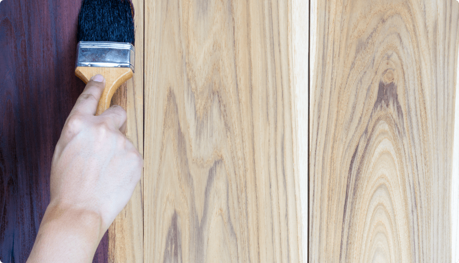 A hand holding a paintbrush applies dark-coloured stain to planks of light-coloured wood.
