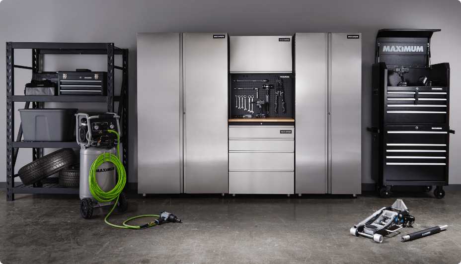 A garage containing an organized storage shelf, a green coiled garden hose on a spool, silver cabinets and drawers, a tidy rack of tools, and car jack on the floor in front of a black MAXIMUM drawer cabinet on caster wheels.