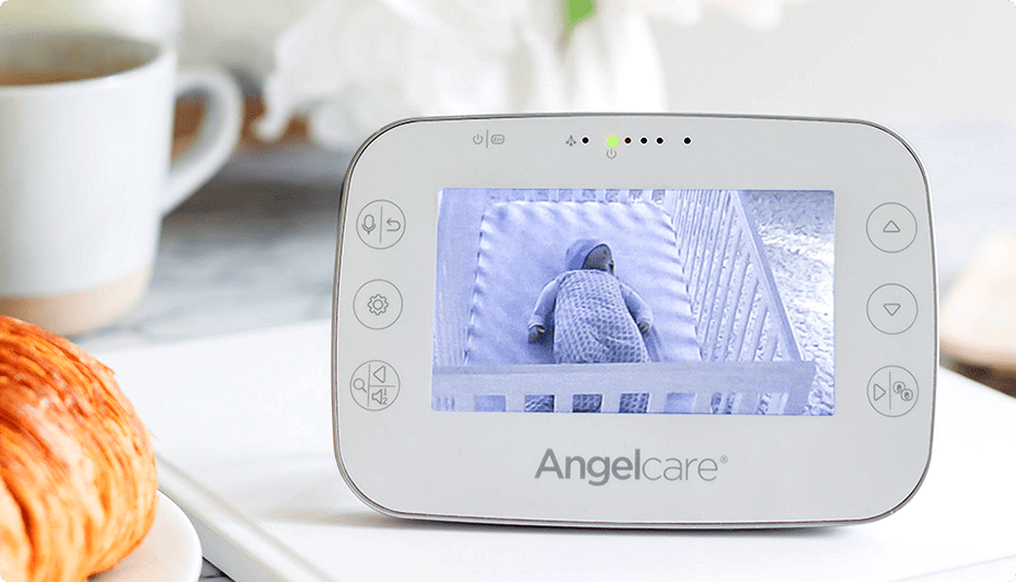 An Angelcare® Baby Breathing Monitor display unit with an image of a sleeping infant on its screen rests on a kitchen counter next to a croissant.