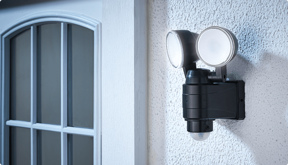 A NOMA 2-Head Security Light mounted on a white stucco wall on the exterior of a home.