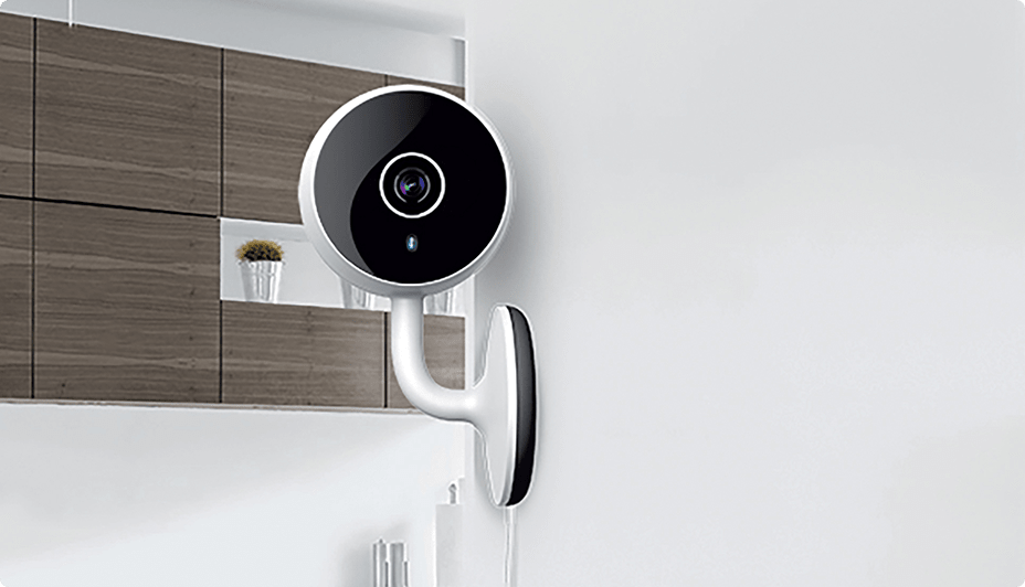 A Geeni HD 1080P Smart Wi-Fi Security Camera mounted on a white wall inside a home.