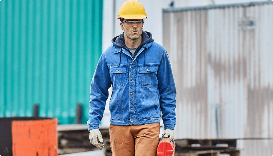 A worker wearing a blue denim jacket, a yellow hard hat, safety glasses, and grey work gloves walks through on a construction job site while carrying a red lunchbox.