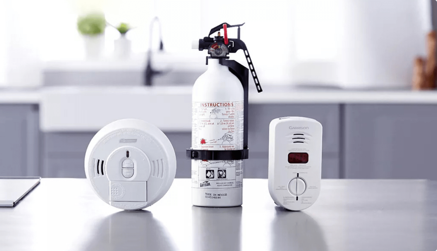 A white smoke detector, fire extinguisher, and carbon monoxide detector rest on a grey countertop inside a home.