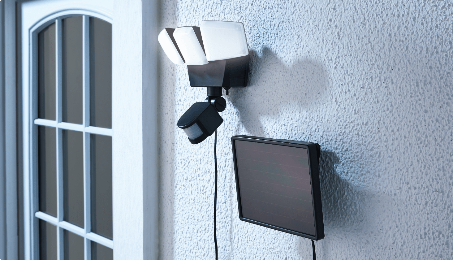 A NOMA 3-Head Security Light and solar panel mounted on a white stucco wall on the exterior of a home.