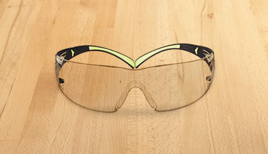 A pair of clear plastic safety glasses with silver frames set against a plywood background.