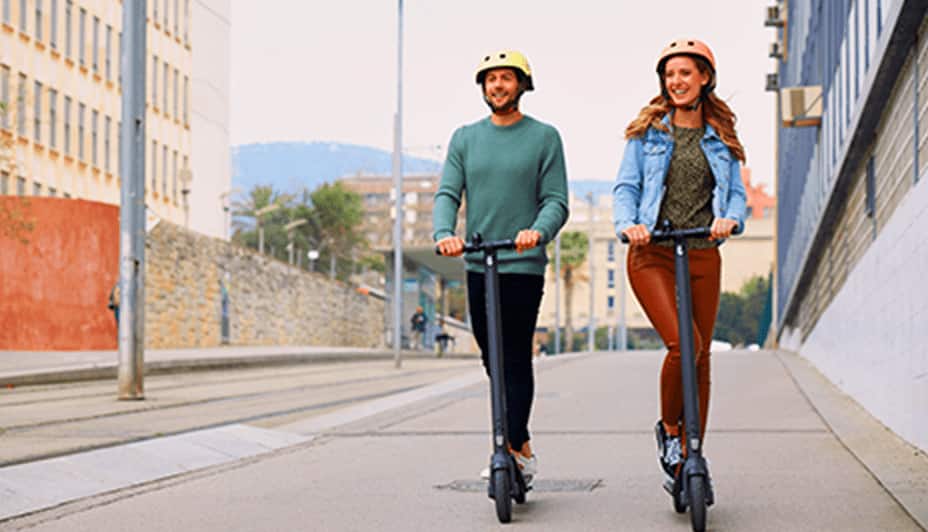 Man and woman in safety helmets riding scooters on a street.