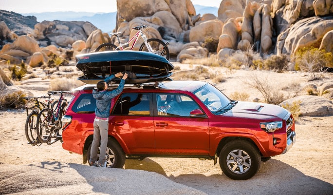 Man uses a rooftop cargo box mounted on a red SUV parked on a mountain road.