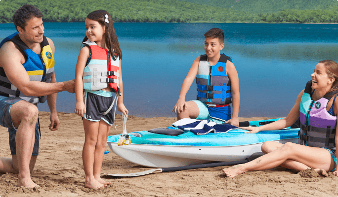 A man, woman, young girl and young boy wearing life jackets gathered around a kayak on a beach. 