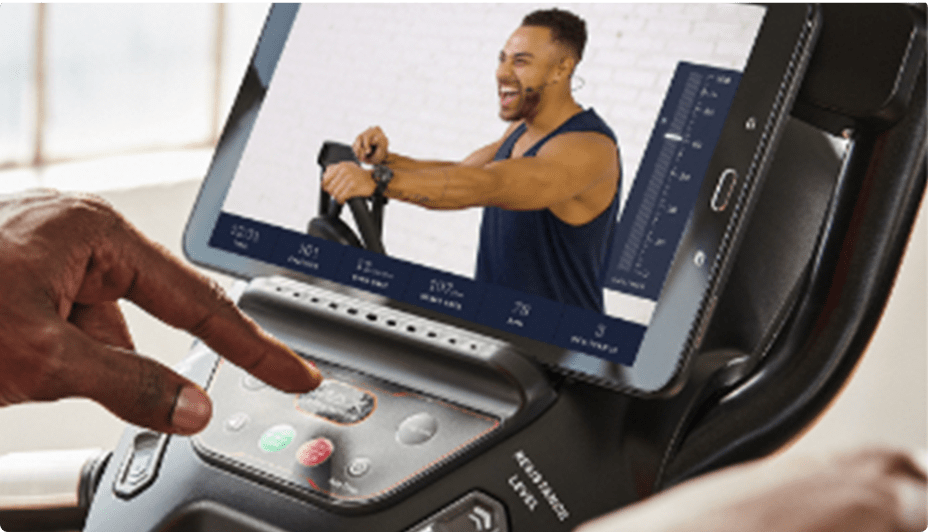 Hand pressing a button on a smart fitness machine with a tablet displaying a workout video.