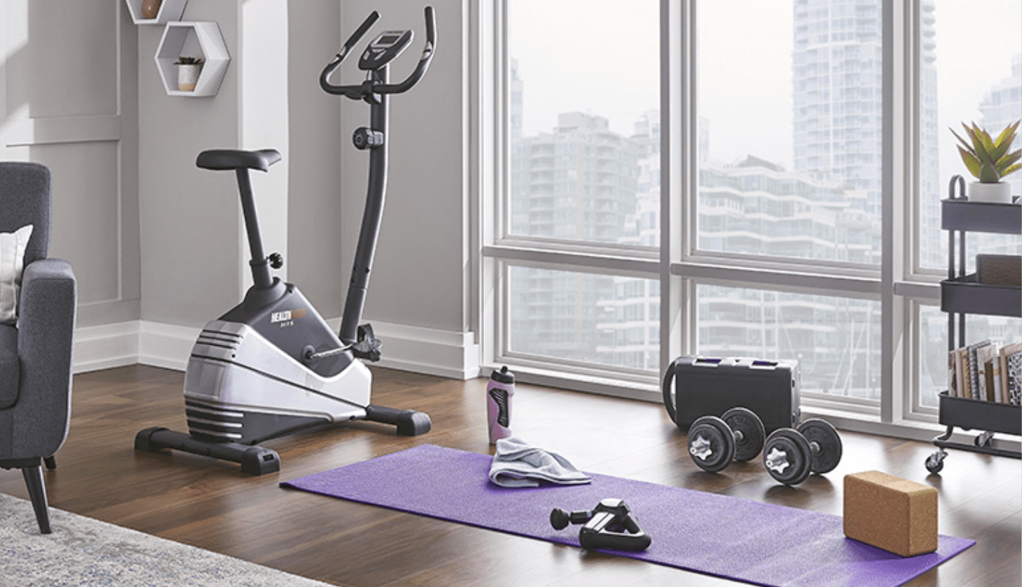 Weights, water bottle, towel on a yoga mat and an exercise bike near a large window. 