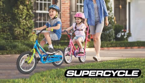 Two young children ride Supercycle bikes in a residential driveway.
