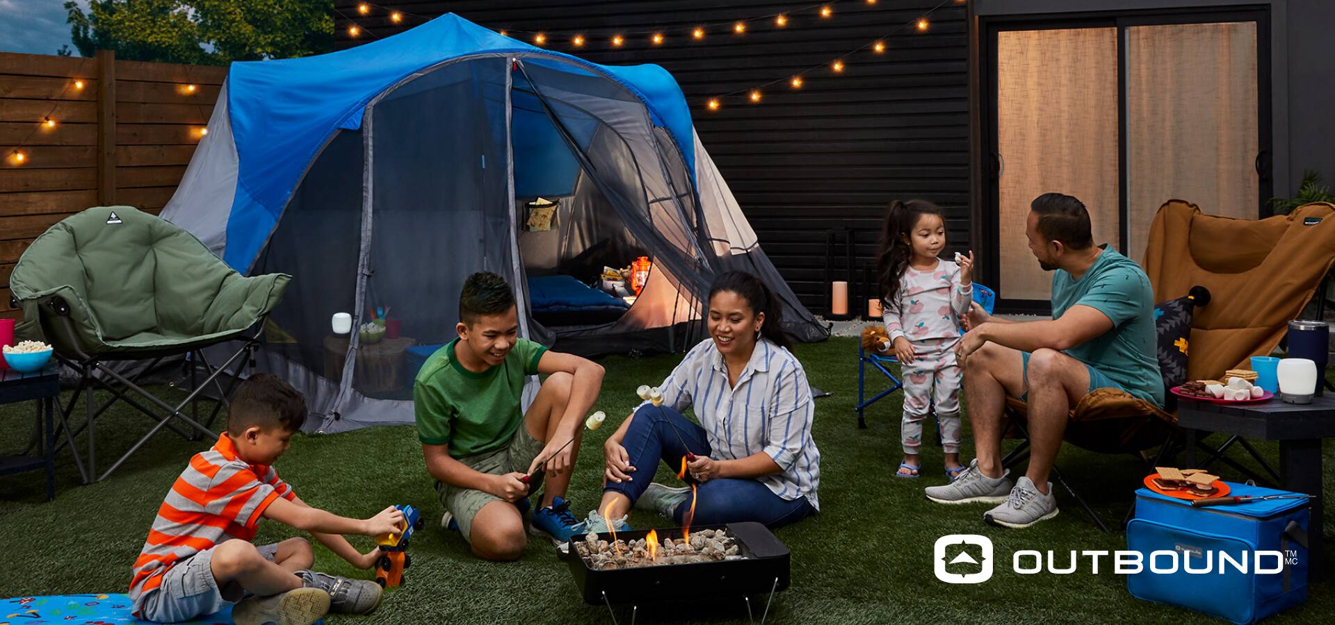 Une famille de deux adultes et trois enfants assis autour d'un feu de camp; une tente et une chaise en arrière-plan.