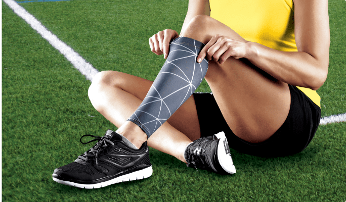 Un athlète assis sur une pelouse artificielle verte remontant un bandage de sport noir sur son genou gauche.