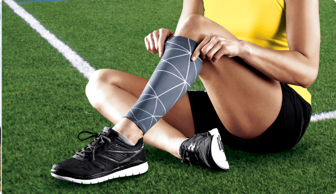 Un athlète assis sur un terrain de sport vert et mettant un manchon de compression pour le mollet Tensor Sport sur sa jambe gauche.