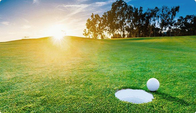 A golf ball lies at the edge of hole on a lush green golf course at sunset.