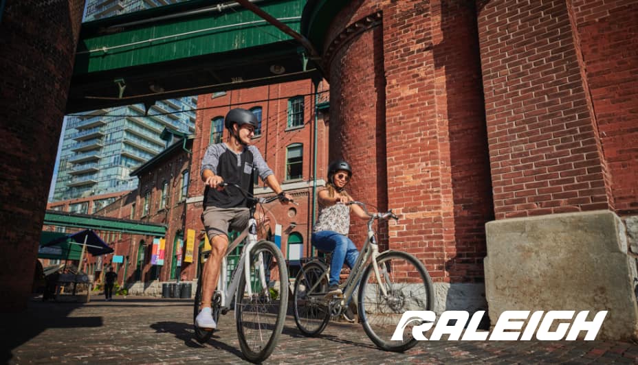 A man and woman riding Raleigh bicycles in a city setting.