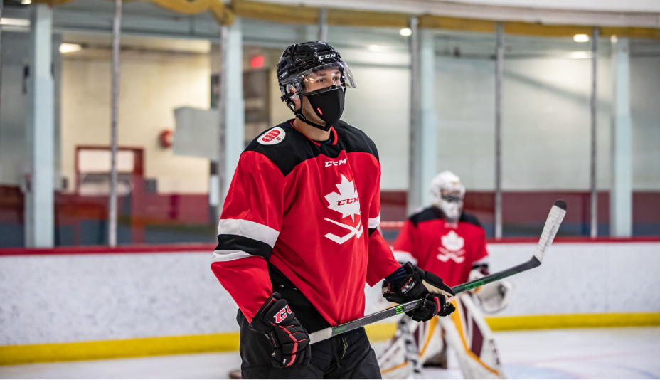 Joueur de hockey adulte portant un masque respiratoire noir et un chandail d’entraînement rouge dans une patinoire de hockey.