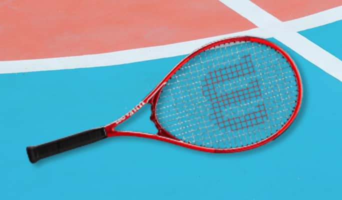 A red Wilson tennis racket lies on a turquoise-and-salmon clay tennis court.