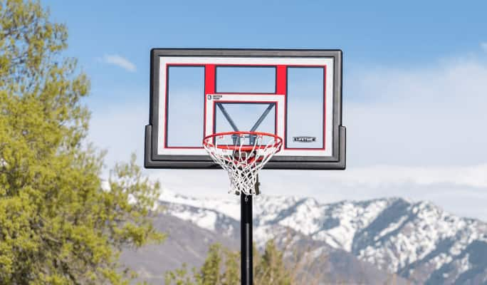 A basketball backboard and rim stand in front of a background of trees, mountains, and blue sky.