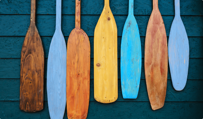 Seven boat and board paddles in natural wood, yellow, and blue.