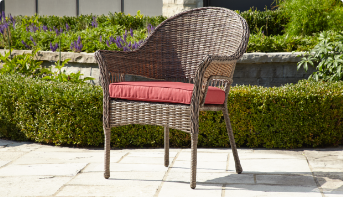 A brown wicker patio armchair with a read cushion placed on it sitting in sunlight. 