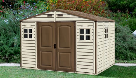 A basic garden shed placed on a lawn with bushes and flowers behind it. 
