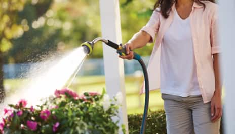 A woman holding out a hose head as it sprays water onto some flowers. 