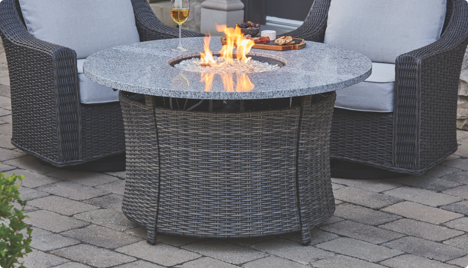 CANVAS Summerhill Round Wicker & Steel Gas Fire Table lit by two chairs.