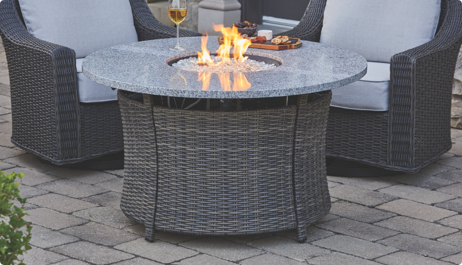 Table ronde de foyer au gaz en osier et acier Summerhill de CANVAS avec un feu allumé et deux chaises.