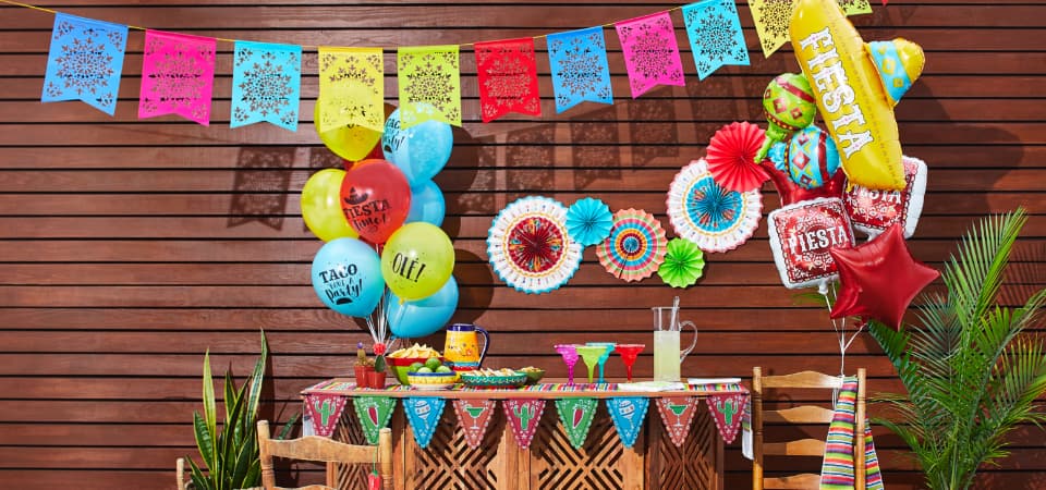 Outdoor party snack table with colourful hanging decorations, and two bouquets of balloons printed with Spanish words including “fiesta.”