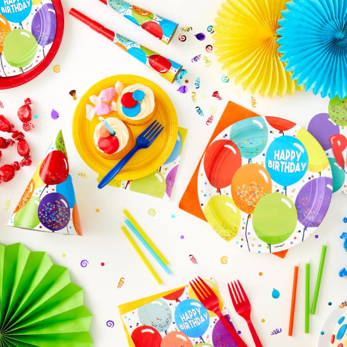 Tabletop covered with colourful birthday-themed tableware, confetti, and paper fans.