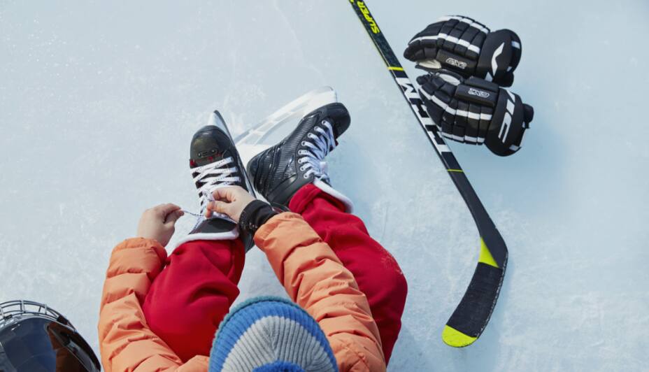 Child putting on skates on ice beside CCM hockey stick