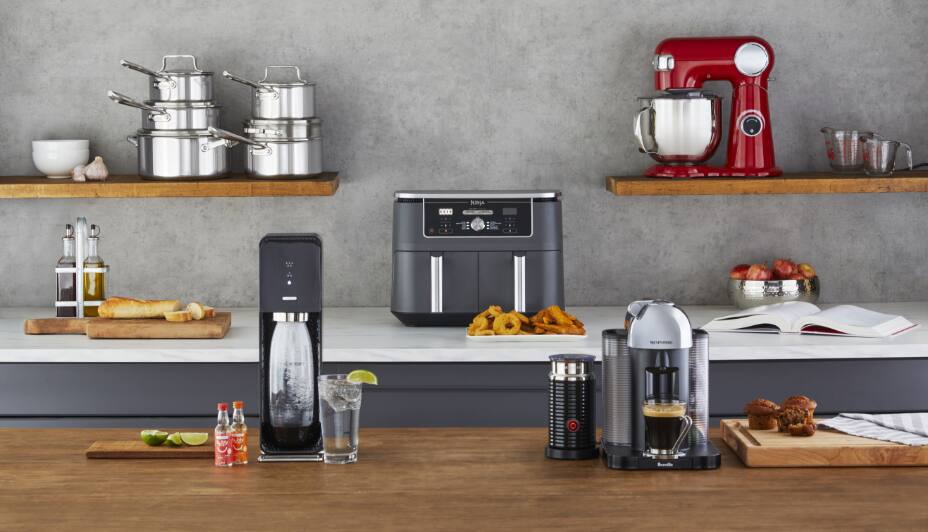 Sélection d'appareils de cuisine, dont une friteuse à air, un appareil SodaStream, un batteur sur socle KitchenAid, une cafetière Nespresso et des casseroles présentés sur un comptoir de cuisine.