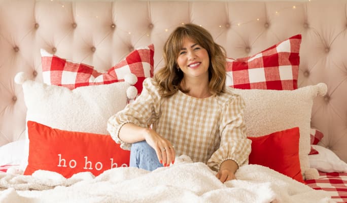 Jillian Harris assise sur un lit décoré de coussins de Noël de sa collection de Noël Jillian Harris x CANVAS