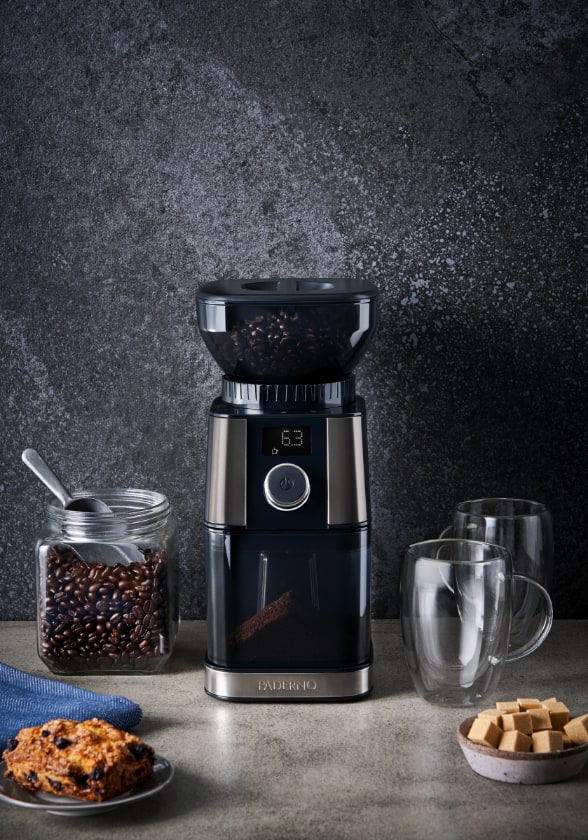 Moulin à café PADERNO avec un contenant de grains de café, des tasses en verre et des pâtisseries à côté