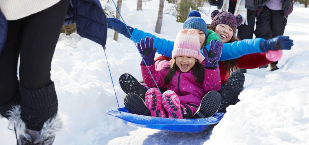 Three overjoyed children wearing winter clothing, hats and boots in snowy outdoor field with teen pulling children on a blue toboggan.