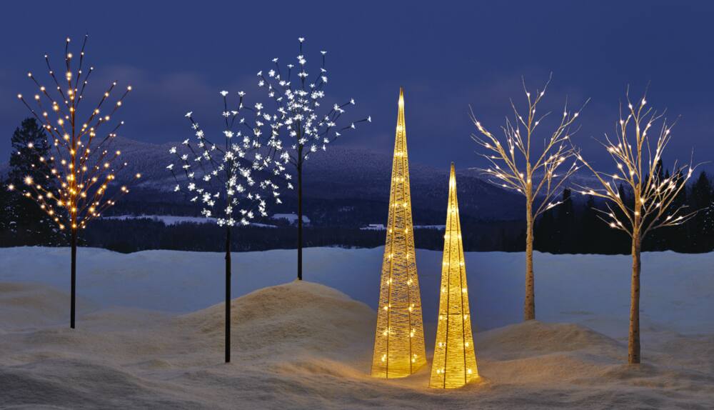 A range of CANVAS Outdoor Pre-Lit Trees on a snowy lawn including the Blossom Tree, Glitter Cone Tree, Birch Tree, and Twig Tree.