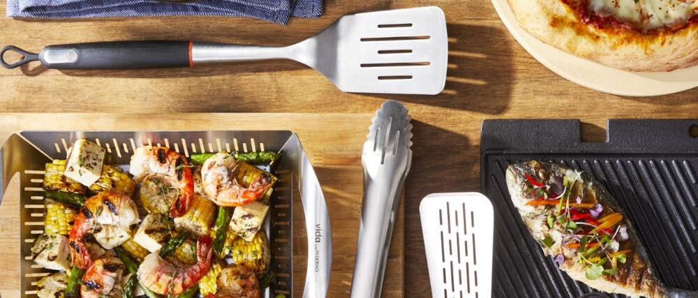 Vida by PADERNO BBQ tongs and spatulas on wooden table with shrimp and fish on grill pans. 