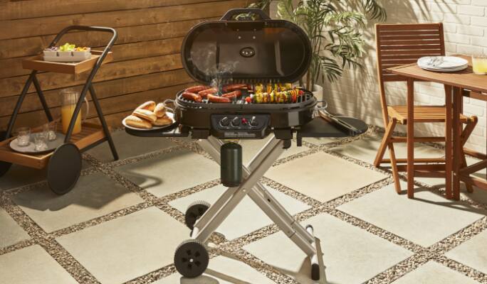 Coleman 3-Burner Roadtrip Cart Grill open in backyard, grilling hot dogs and skewers. 