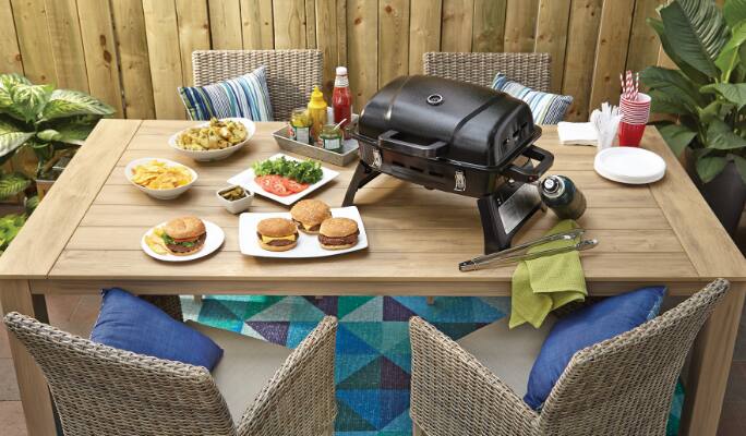 Barbecue portatif au gaz MASTER Chef sur une table à manger extérieure avec hamburgers et légumes dans des assiettes.