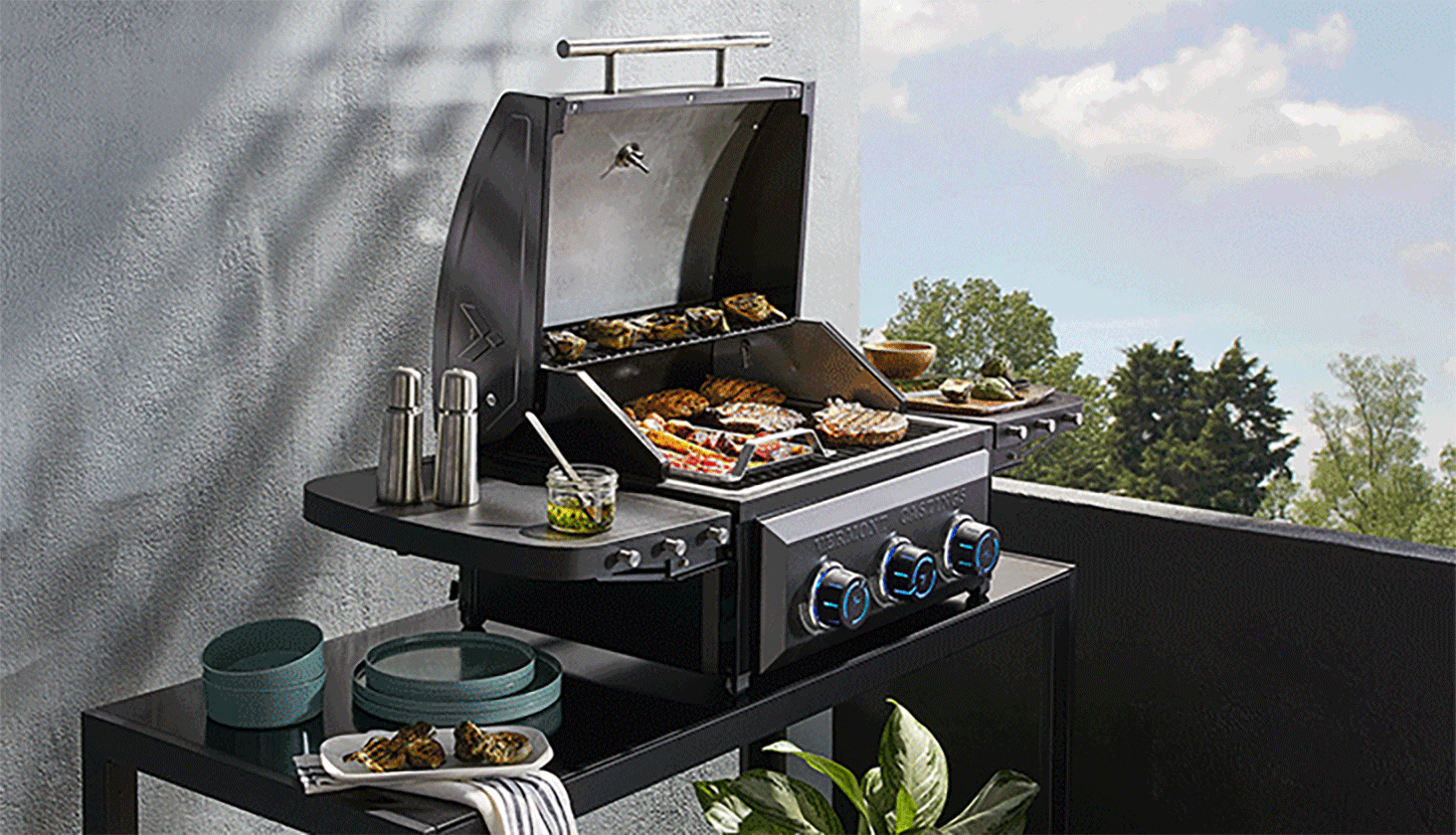 Vermont Castings Ascent 3-Burner Electric Balcony Grill on outdoor table with side panels open, grilling burgers.