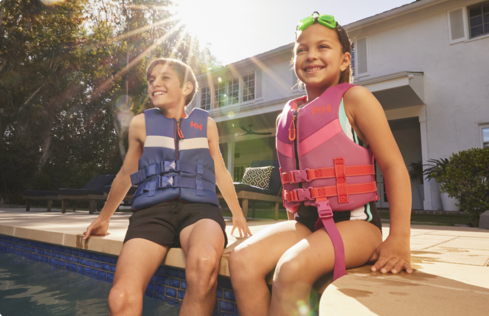 Two kids sitting by pool wearing life jackets 