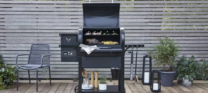 A black Coleman Cookout™ Pellet Grill set against a brown horizontal-slat fence in a backyard.