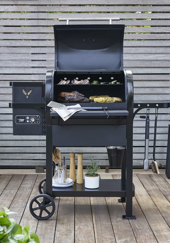 A black Coleman Cookout™ Pellet Grill set against a brown horizontal-slat fence in a backyard.
