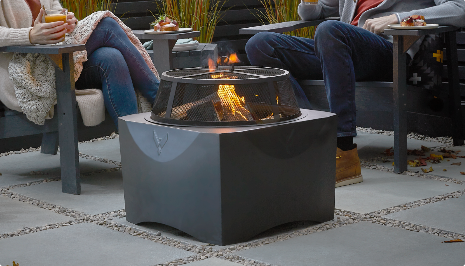 Two people relax in comfortable chairs around a lit fire bowl in a backyard at twilight.