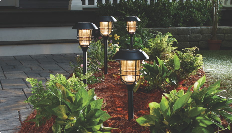 NOMA Solar Flame Torch Stake Lights are displayed on a front porch garden with plants.