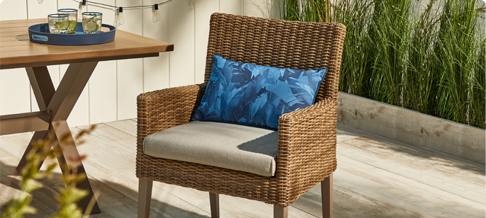 CANVAS Belwood Dining Chair on wooden backyard porch dining area featuring plush cushion seat, decorative tropical pillow and wicker accents on the chair.